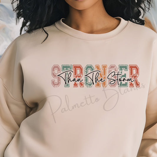 Beige sweatshirt with “Stronger Than The Storm” design in distressed multicolor varsity block letters and script overlay, ideal for fall screen print transfers.