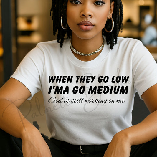 Screen print transfer design featuring the phrase “When They Go Low I’ma Go Medium – God is still working on me” in bold black lettering on a white shirt worn by a woman.